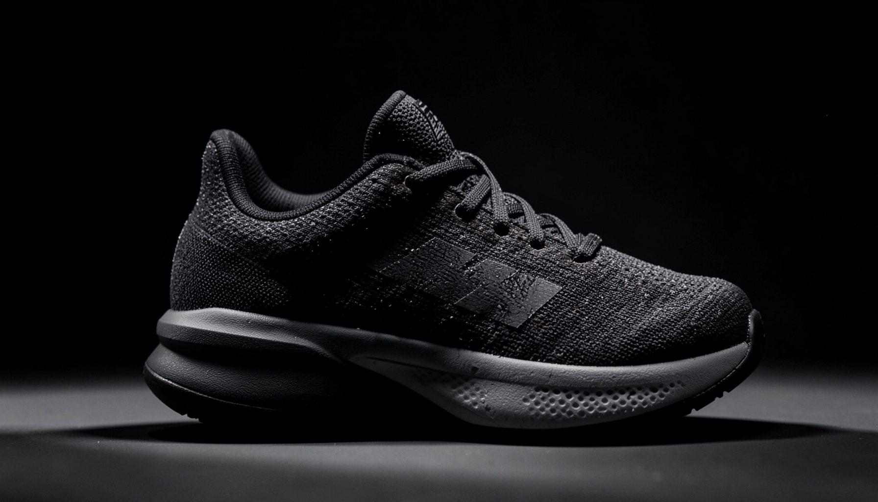 Dark-toned adidas basketball shoe with textured upper, dramatic side lighting on a shadowed backdrop