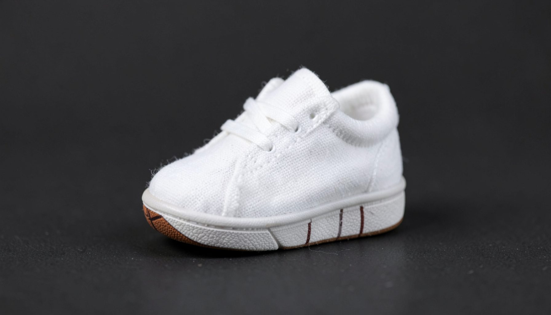 Tiny infant sneaker in soft white cotton with a miniature basketball-inspired sole, photographed on a neutral dark background