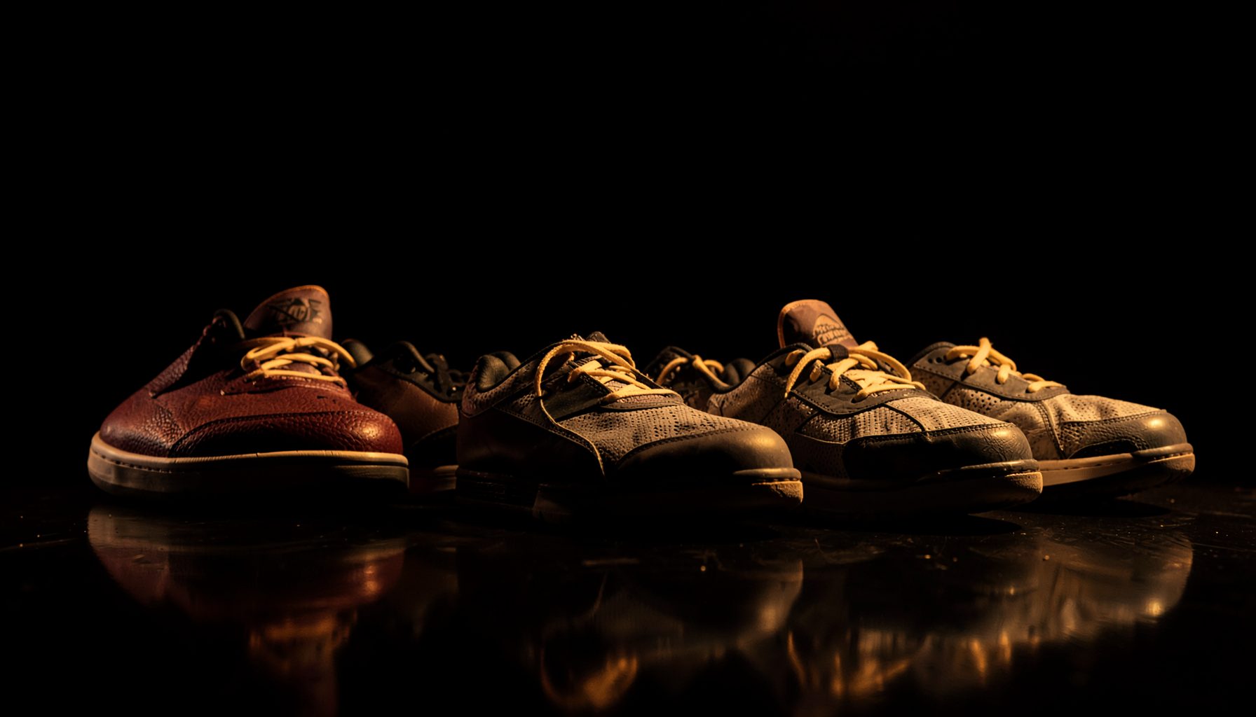 Dark atmospheric display of basketball sneakers lined up on a glossy floor, with dramatic shadows and warm amber accent lighting against a black background