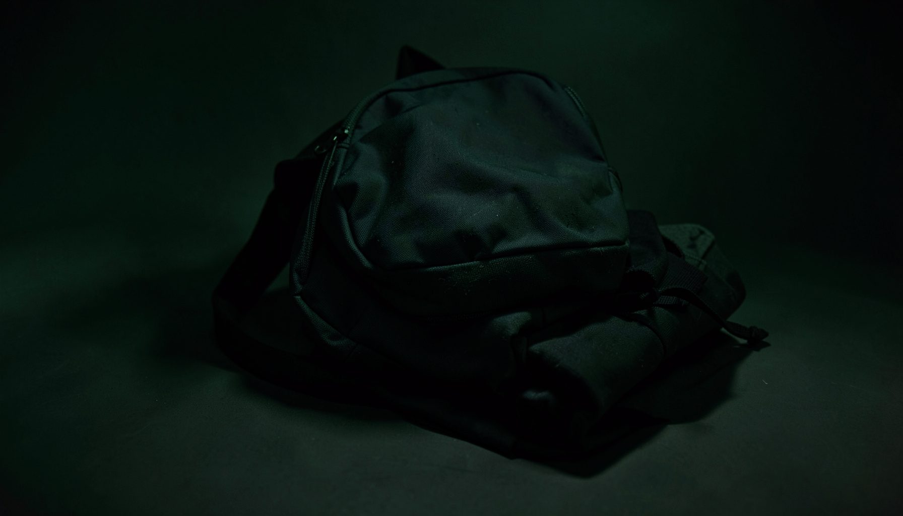 Dark backpack and folded apparel on a shadowed surface, subtle green accent lighting